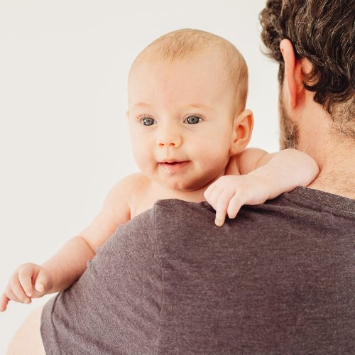 Young happy baby peaks over his dads shoulder.